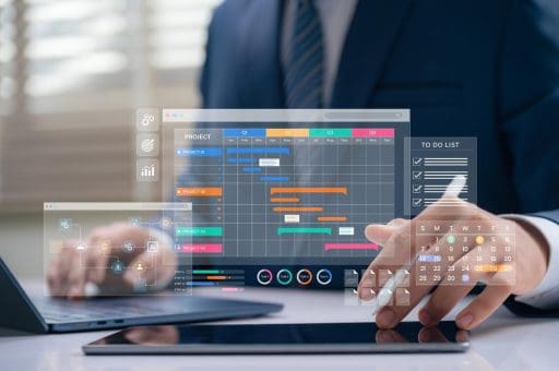 A man in a suit sat at a desk in front of a laptop. The screen is reflected int he glass displaying a project plan and to do list.