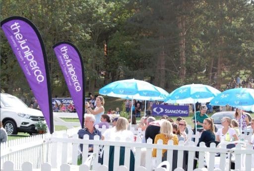 Attendees sitting outdoors around round tables with blue parasols . With two The Juniper Co purple banners on the outside of the area