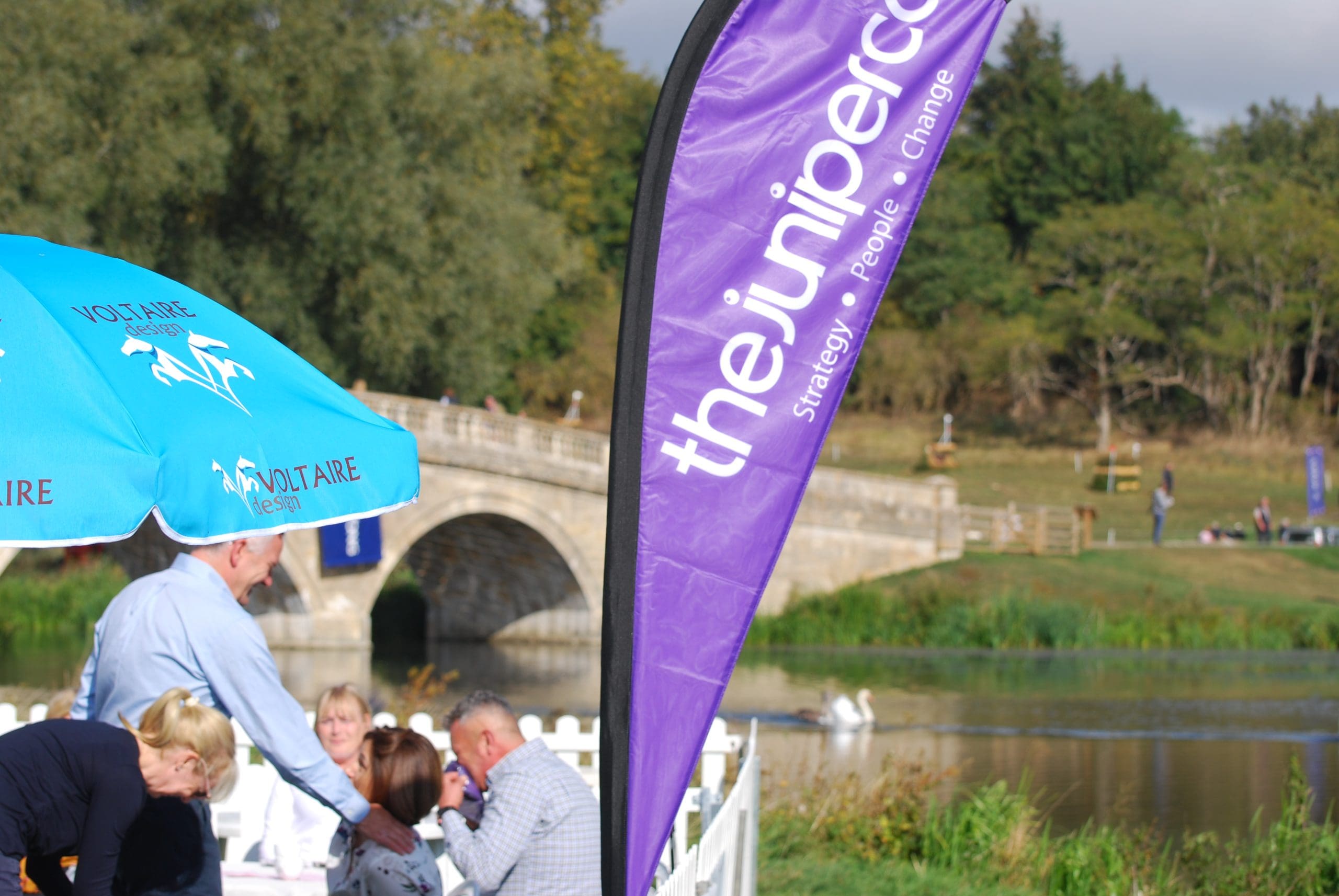 A purple feather flag with The Juniper Co. branding, in front of the lake at Blenheim Horse Trials. People sit behind in a white picket fence enclosure.