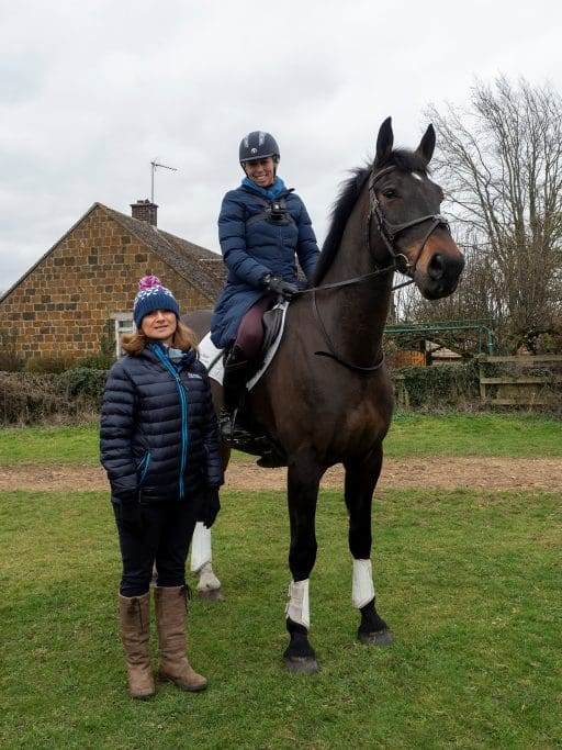 Nicki Kavanagh stands next to Ginny Howe who is seated on a horse in a field .