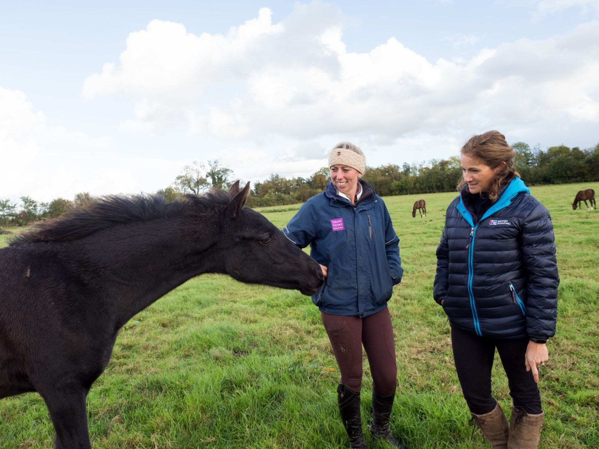 Ginny Howe and Nicki Kavanaugh standing in a field next to a black horse. There are two horses in the field behind.