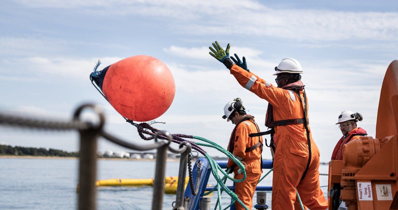 A photograph of a team of people working on an oil rig out at see, wearing personal protective equipment. A man in the foreground is throwing an orange buoy into the water.