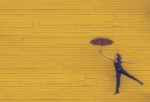 Woman holding an unfolded umbrella in her outstretched left hand and jumping in front of a yellow background